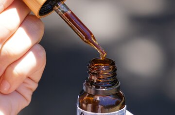 A dropper full of dark liquid hovers above a small brown bottle 