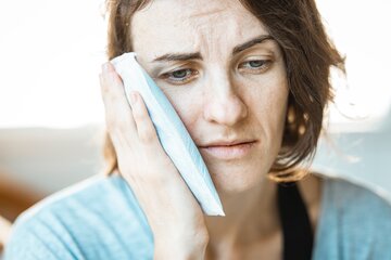 A woman holds an ice pack to her face with an expression of discomfort.