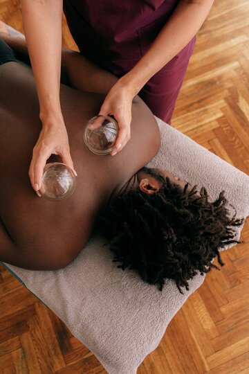 A man with short dreads lays facedown while receiving a cupping treatment from a person in maroon scrubs.